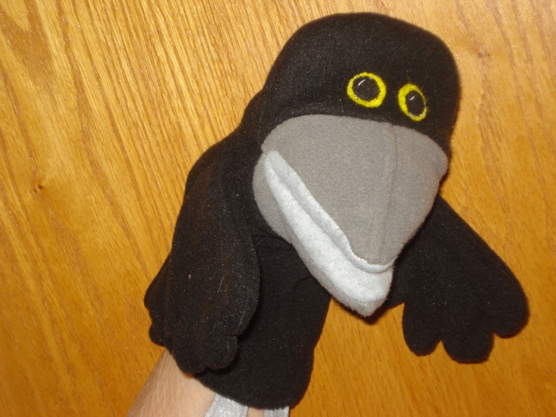 May include: A black crow hand puppet made of soft fabric. The bird features a large grey beak, a light grey lower jaw, and two round yellow eyes with black pupils. The puppet has two small black wings extending from the sides of its body.