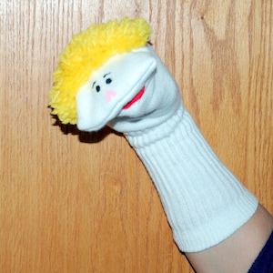 May include: A handmade sock puppet with a white face, yellow hair, and a red mouth. The puppet has black eyes and pink cheeks. The sock is white and ribbed, with a person's arm inside.