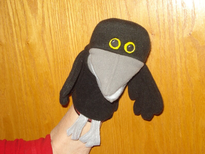 May include: A black bird hand puppet featuring a large grey beak, yellow eyes with black pupils, and two grey feet dangling from the bottom. The puppet is constructed from soft black fabric with stitched details on the wings and head.