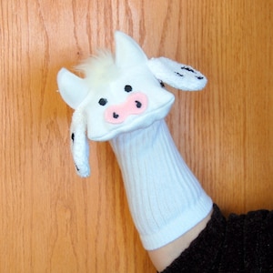 May include: White sock puppet with a cow face. The puppet has black eyes, a pink nose, and black spots on the ears.