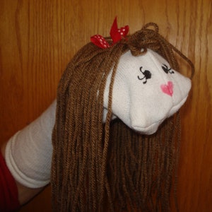 May include: A handmade sock puppet with a white head, brown yarn hair, and a red bow with white polka dots. The puppet's face has black eyes and a pink heart-shaped mouth. The puppet is on a white sock arm.