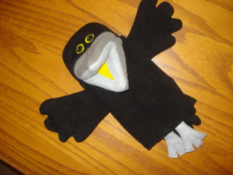 May include: A black bird hand puppet featuring a grey beak with a yellow interior, yellow eyes with black pupils, and light grey tail feathers. The puppet has outstretched wings and a rounded head, constructed from soft fabric material.