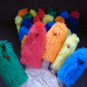 May include: A set of colorful finger puppets made of fuzzy fabric. The puppets are in various colors, including red, orange, yellow, green, blue, and purple. Each puppet has a simple face with eyes and a mouth.