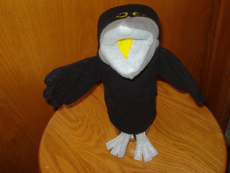 May include: A black bird hand puppet made of soft fleece fabric. The puppet features a light grey face, a bright yellow beak, and two yellow eyes. It has outstretched wings and light grey feet at the base, designed for interactive play.