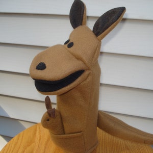May include: A brown kangaroo puppet with black ears and a black mouth. The puppet has a small brown joey in its pouch.