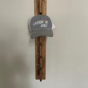 May include: A wooden hat rack with a grey and white Landshark Golf hat hanging on it. The rack is made of dark stained wood and has a simple design. The hat has the words "LANDSHARK GOLF" embroidered on the front.