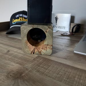 May include: A wooden phone stand with a circular aperture, holding a black smartphone. The stand is made from a square block of wood with visible grain and a natural finish. A black hat with the text "Live the Game LandSha" is visible in the background.