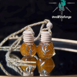 May include: Two small, clear glass bottles filled with a shimmering, golden liquid. Each bottle has a wooden cap and a silver charm featuring a crescent moon and a cat. The bottles are suspended by a rope and are reflected on a dark surface.