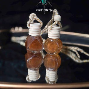 Puede incluir: Dos pequeñas botellas de vidrio transparente con líquido marrón y tapas de madera blanca suspendidas por una cuerda. Las botellas se reflejan en una superficie oscura y reflectante. El texto "@veilfireforge" es visible.