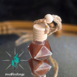 Op de afbeelding: Een kleine, geometrisch gevormde glazen fles gevuld met een bruine vloeistof, afgewerkt met een houten dop en een decoratief touw. De fles hangt aan een touw met een houten kraal. Het logo @veilfireforge is zichtbaar.