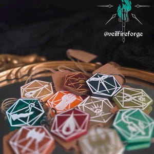May include: A collection of colorful, geometric pendants with various symbols and designs. Each pendant is a different color, including green, orange, and red, and is attached to a small tag. The image also features the text "@veilfireforge".