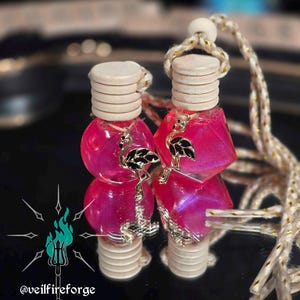 May include: Two pink, decorative car air fresheners with silver flamingo charms. Each bottle has a wooden cap and a decorative cord. The image includes the text "@veilfireforge".