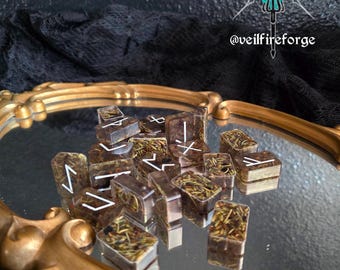 May include: A collection of rectangular, translucent resin runes with white markings and dried herbs inside. The runes are displayed on a mirrored surface within a gold-colored, ornate frame. The background is dark, with the text "@veilfireforge".
