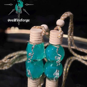 May include: Two teal-colored car air fresheners with wooden caps and silver accents. Each freshener is suspended by a beige and gold cord. The background is dark, with the text "@veilfireforge" and a logo.