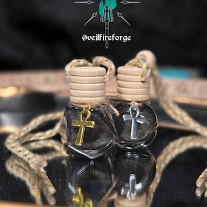May include: Two small, clear glass bottles filled with dark liquid, each topped with a wooden cap and a decorative ankh charm. One charm is gold, the other silver. The bottles hang from a beige rope. The text "@veilfireforge" is visible.
