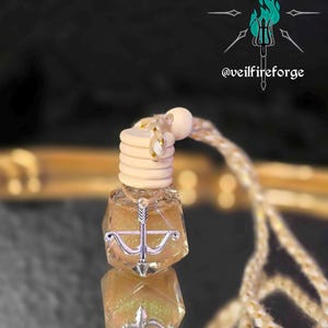 May include: A small, clear glass bottle filled with a shimmering, golden liquid. The bottle has a wooden cap and a silver-colored charm in the shape of a bow and arrow. A decorative cord is attached to the bottle. The text "@veilfireforge" is visible.