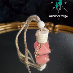 May include: A pink, geometric-shaped car air freshener with a gold-colored key charm. The air freshener hangs from a beige and gold-speckled cord with a wooden bead. The background is a reflective surface with the text "@veilfireforge".