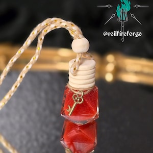 May include: A small, clear glass bottle filled with red liquid and a gold-colored key charm. The bottle has a wooden cap and is suspended by a beige and gold rope. The image includes the text "@veilfireforge".