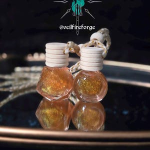 May include: Two small, clear glass bottles filled with shimmering gold liquid, topped with light-colored wooden caps and a woven cord. The bottles are reflected on a dark, reflective surface. The image includes a logo with text that reads "@veilfireforge".