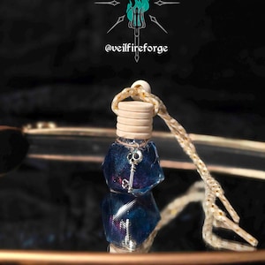 May include: A small, blue, geometric-shaped bottle with a silver key charm hanging from a wooden stopper. The bottle is suspended by a beige and gold rope. The text "@veilfireforge" is visible in the background.