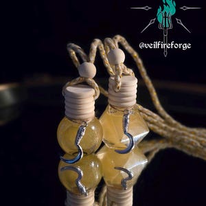 May include: Two glass car air fresheners with a yellow liquid and silver crescent moon charms. The wooden tops are connected by a rope with gold accents. The background is dark with the text "@veilfireforge".