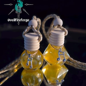 May include: Two glass car air fresheners with yellow liquid inside. Each has a wooden cap, a silver musical note charm, and a rope for hanging. The background is dark, and the logo "@veilfireforge" is visible.
