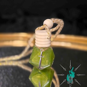 May include: A green, geometric-shaped car air freshener with a silver sword charm. The air freshener has a wooden top and a beige rope for hanging. The background is dark and reflective.