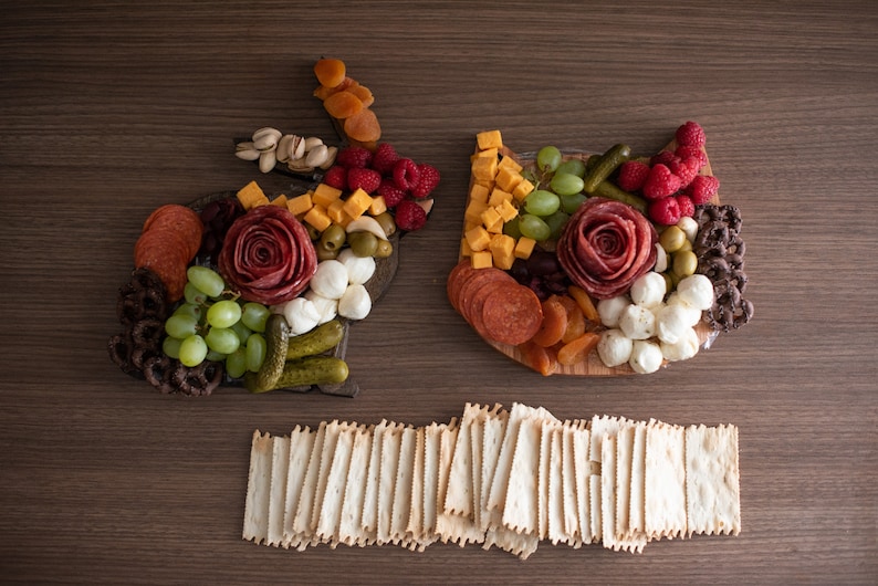 May include: A charcuterie board shaped like a bunny and a cat, with a variety of meats, cheeses, fruits, and vegetables. The board is surrounded by a pile of crackers.
