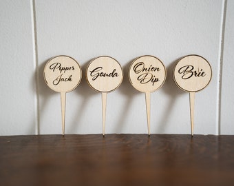 May include: Four wooden round signs with black text on sticks. The signs read "Pepper Jack", "Gouda", "Onion Dip", and "Brie".