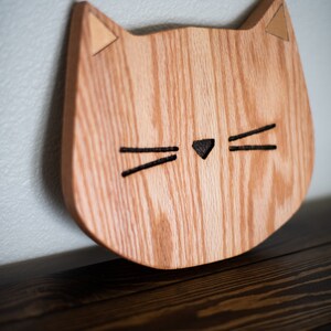May include: A wooden cat-shaped wall decor with a light brown wood grain and a black cutout for the eyes and nose. The ears are made of a lighter wood and are triangular in shape.