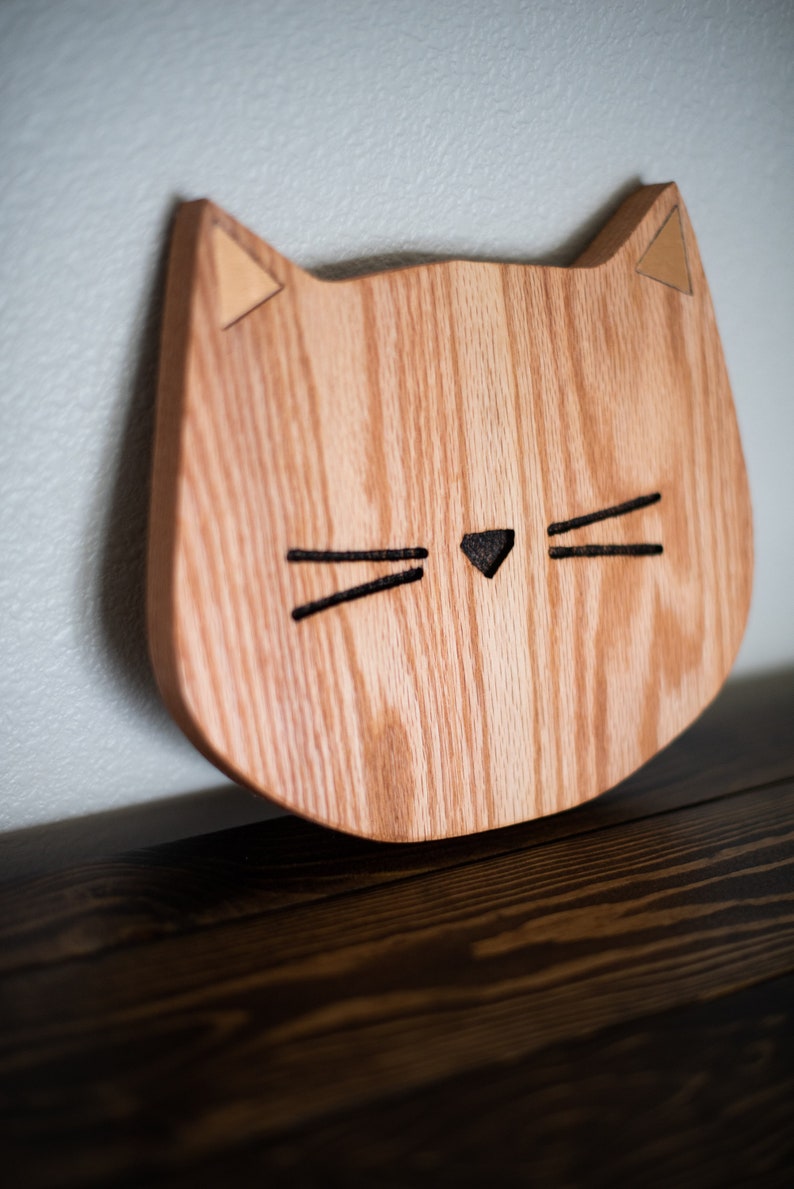 May include: A wooden cat-shaped wall decor with a light brown wood grain and a black cutout for the eyes and nose. The ears are made of a lighter wood and are triangular in shape.