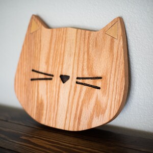 May include: A wooden cat head wall decor with a light brown wood grain and gold ears. The cat has black whiskers and a black nose.