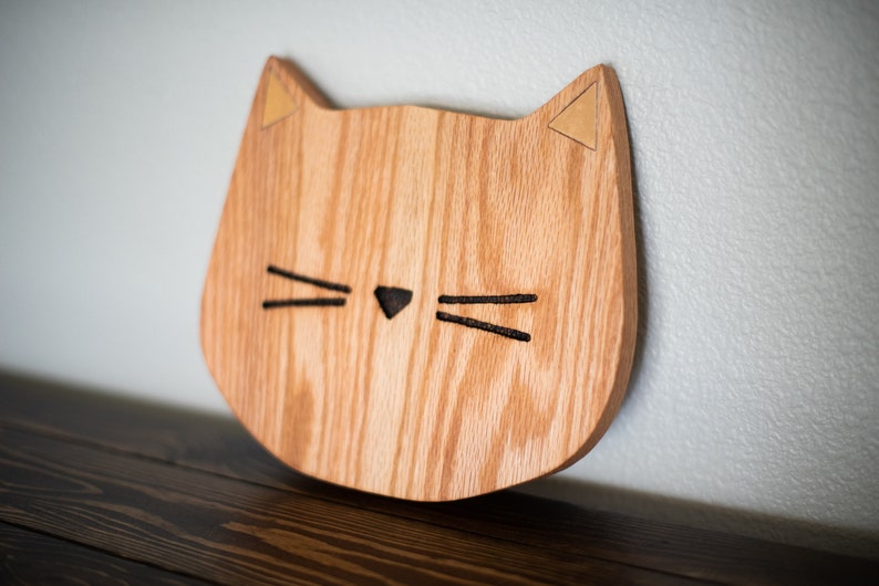 May include: A wooden cat head wall decor with a light brown wood grain and gold ears. The cat has black whiskers and a black nose.