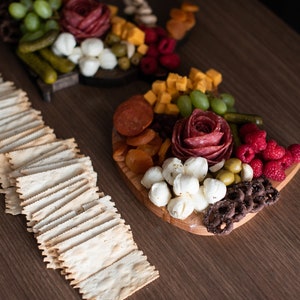May include: A wooden serving board with a variety of snacks, including salami, cheese, grapes, pretzels, and crackers. The board is arranged in a decorative pattern.