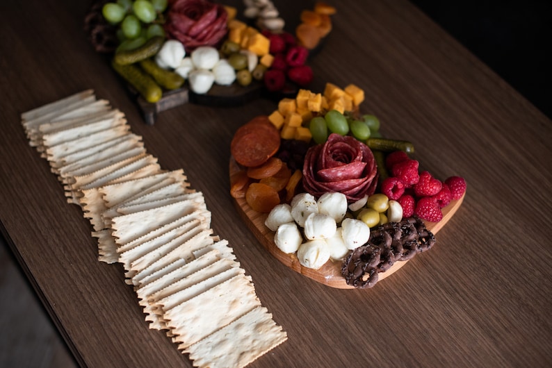 May include: A wooden serving board with a variety of snacks, including salami, cheese, grapes, pretzels, and crackers. The board is arranged in a decorative pattern.