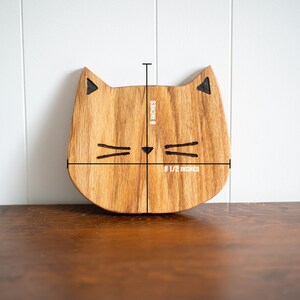 May include: Wooden cat-shaped cutting board with a natural wood finish. The board measures 8 1/2 inches wide and 8 inches tall.