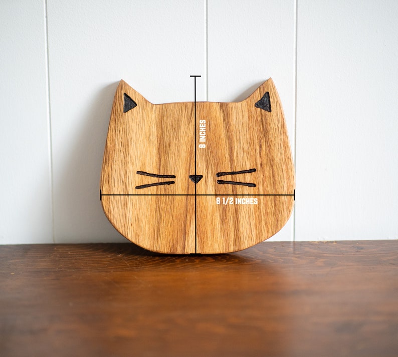 May include: Wooden cat-shaped cutting board with a natural wood finish. The board measures 8 1/2 inches wide and 8 inches tall.