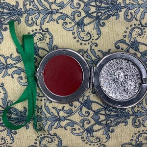 May include: A silver-colored, round locket with a hinged design, open to reveal a deep red circle on one side and an ornate, engraved pattern on the other. A green ribbon is attached for hanging.