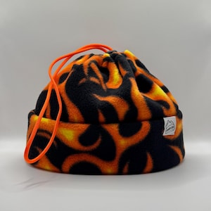 May include: A fleece beanie featuring a black and orange flame pattern. The hat has a folded brim and a bright orange drawstring cord at the top. A small white rectangular tag with a black line drawing of a mountain and text is attached to the side.