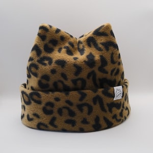 May include: A tan and black leopard print beanie with a folded cuff and two pointed ears at the top. A small white rectangular tag is sewn onto the side of the cuff, featuring a black line drawing of a mountain and the text "MOUNTAIN OPS".