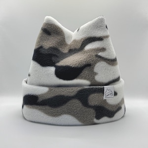 May include: A fleece beanie featuring a white, grey, and black camouflage pattern with two pointed ears at the top. The hat has a folded cuff and a small white rectangular fabric tag on the side with the text WONDER CAPS and a mountain icon.