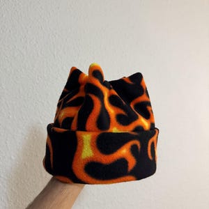 May include: A black, orange, and yellow flame-patterned beanie hat. The hat is made of a soft, fleece-like material. The beanie has a folded brim and a crown with a unique, pointed design.