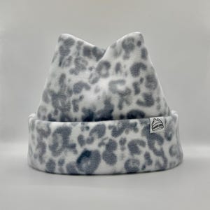 Puede incluir: Un gorro de forro polar con un estampado de leopardo en blanco y gris. El gorro presenta dos orejas puntiagudas en la parte superior y un borde doblado. Una pequeña etiqueta de tela rectangular blanca está cosida en el borde, con el texto PONDER CAPS.