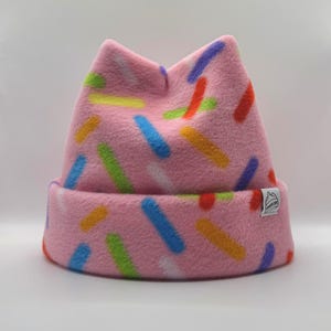 May include: A pink fleece beanie with cat-ear shaped points at the top. The hat features a colorful pattern of short, horizontal dashes in blue, orange, green, purple, and white. A small white rectangular tag is sewn onto the folded brim.