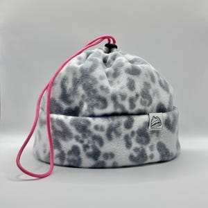 Può includere: Un berretto in pile con un motivo animalier bianco e grigio. Il cappello include una coulisse rosa brillante con un fermacorda nero sulla parte superiore per la regolazione. Una piccola etichetta rettangolare bianca sul risvolto mostra un logo a forma di montagna e il testo POWDER CAPS.