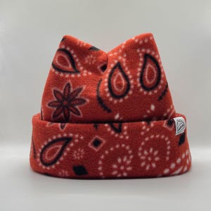 May include: A bright orange fleece beanie featuring a black and white paisley pattern. The hat has a folded cuff and a small white rectangular tag on the side with a black graphic logo of a mountain peak and the text "SUMMER CAPS".