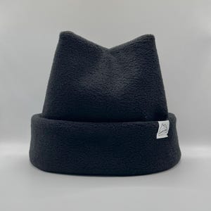 May include: A dark charcoal gray fleece beanie with a folded cuff and a pointed top. A small white rectangular fabric tag is sewn onto the side of the cuff, featuring a black line drawing of a mountain range and the text "SUMMIT CAPS".