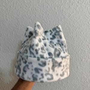 Könnte beinhalten: Eine weiße und graue Fleece-Mütze mit Leopardenmuster. Die Mütze hat einen umgeschlagenen Rand und eine kronenartige Form. Die Mütze ist aus einem weichen, plüschigen Material gefertigt.