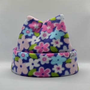 May include: A fleece beanie with a dark blue background featuring a repeating floral pattern in shades of pink, light blue, and white, accented with green leaves. The hat has a folded brim and a small white rectangular tag with black text.