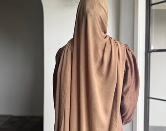 Op de afbeelding: Een persoon die een bruine hijab en een jurk met lange mouwen draagt. De hijab is over het hoofd en de schouders gedrapeerd en de jurk heeft lange mouwen. De outfit is effen van kleur, met een neutraal palet.